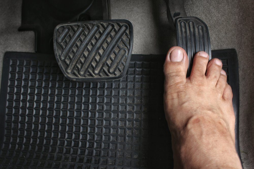 A bare foot presses on a car pedal while resting on a textured mat. The vehicle's interior is visible, indicating a driving context.