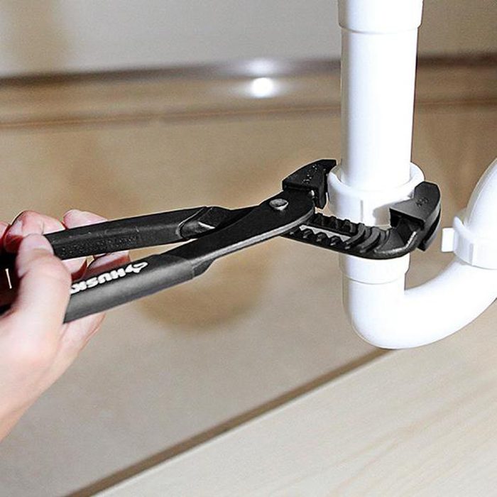 A hand grips a pair of pliers, adjusting a white plumbing pipe under a sink, with a light-colored surface in the background.