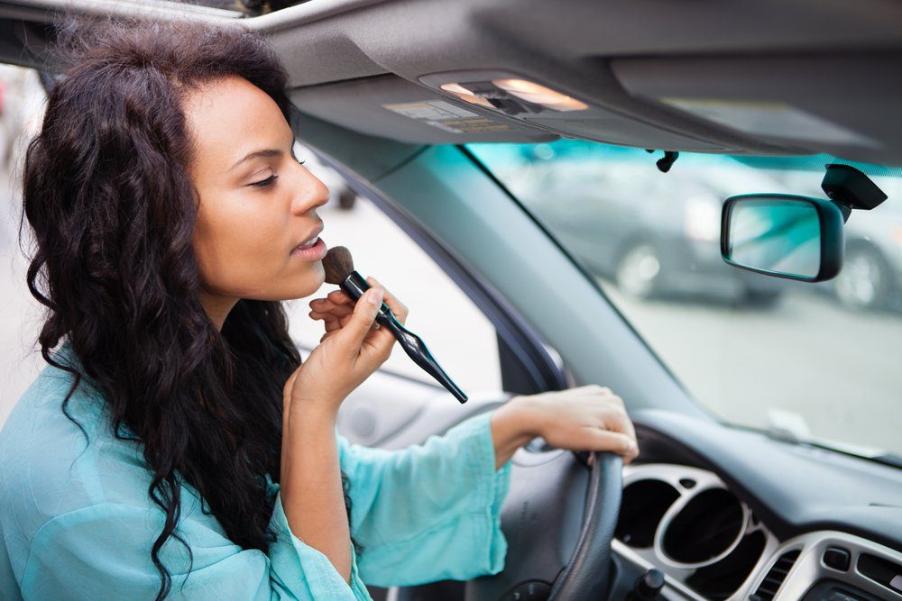 A woman applies makeup with a brush while sitting in a car, focusing on her reflection in the mirror, surrounded by blurred vehicles outside.