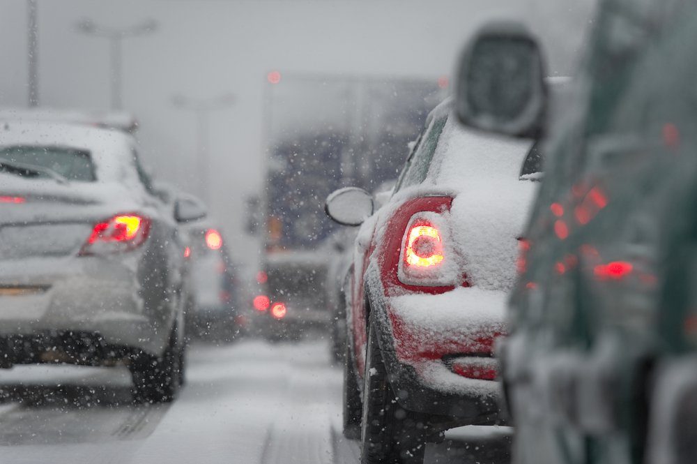 Cars are stuck in traffic, covered in snow, as heavy snowfall reduces visibility on a wintery road. Brake lights glow softly in the distance.