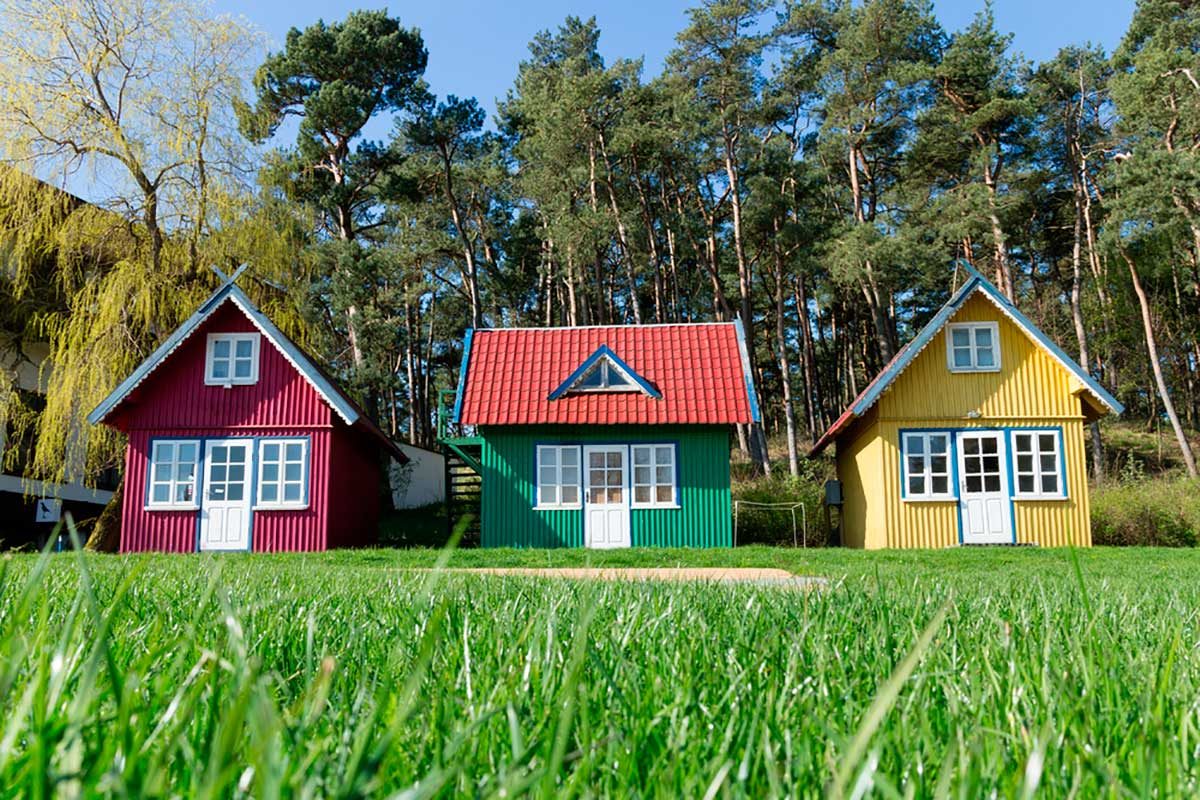 Three colorful houses—red, green, and yellow—stand on vibrant green grass, surrounded by tall pine trees under a clear blue sky.