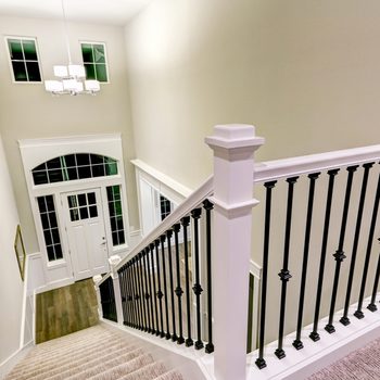 A staircase with a white rail and black spindles descends into a well-lit foyer, featuring a glass-paneled front door and chandeliers above.