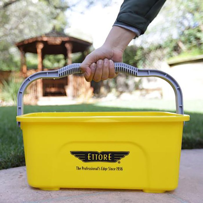 A hand holds a yellow cleaning bucket with a gray handle, set on a stone path. A gazebo and greenery are blurred in the background.