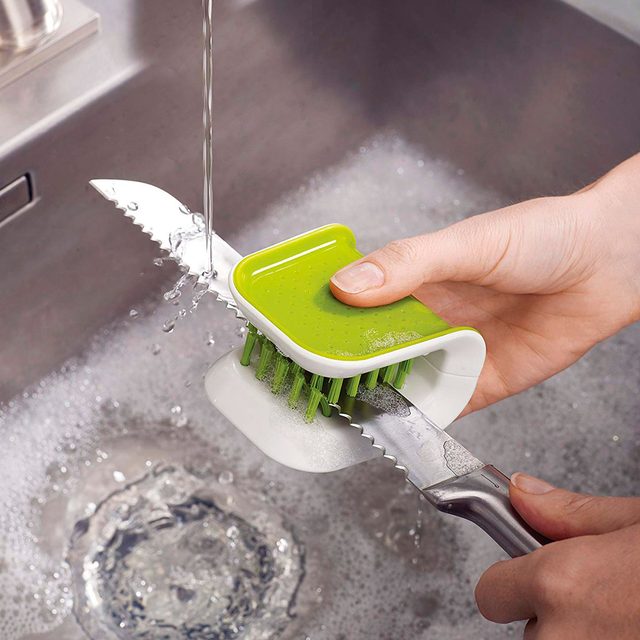 A hand holds a knife while brushing it with a cleaning tool under running water in a sink filled with bubbles.