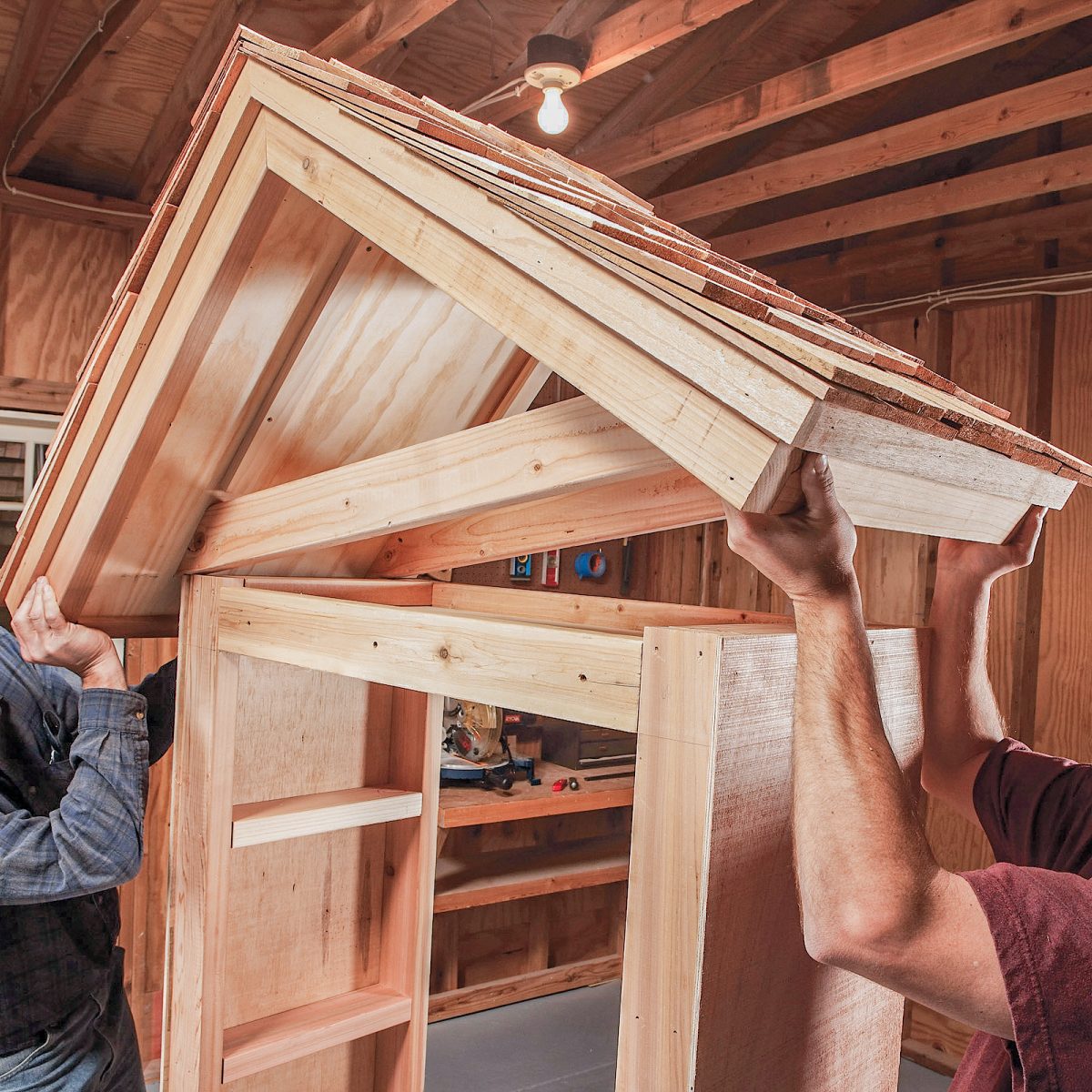 Two individuals are lifting a wooden roof structure, assembling it onto a wooden frame inside a workshop with visible tools and wooden walls.