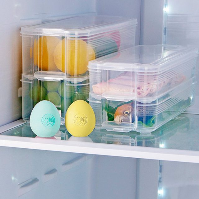 Transparent containers filled with colorful fruits and vegetables sit in a refrigerator, while two decorative eggs rest on the glass shelf beside them.