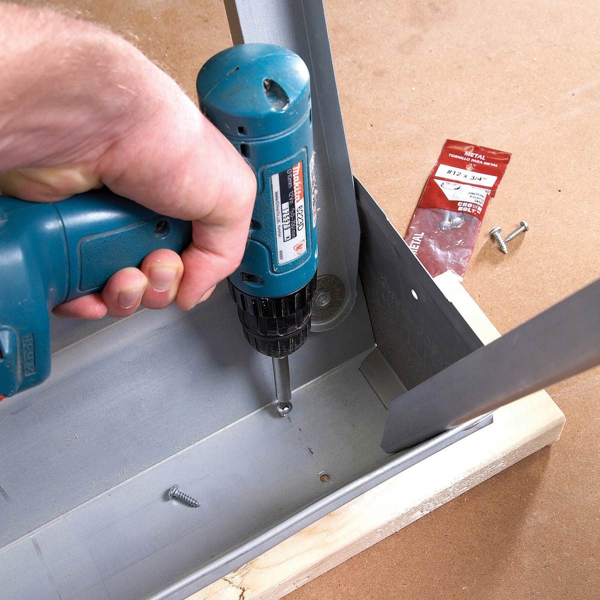 A hand holds a drill to fasten screws into a metal object, resting on a wooden surface, with additional screws and packaging nearby.