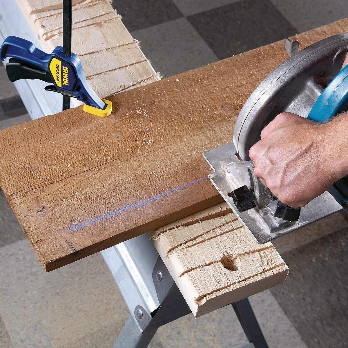A hand uses a circular saw to cut a wooden board, secured with a clamp, on a workbench surrounded by a workshop environment.
