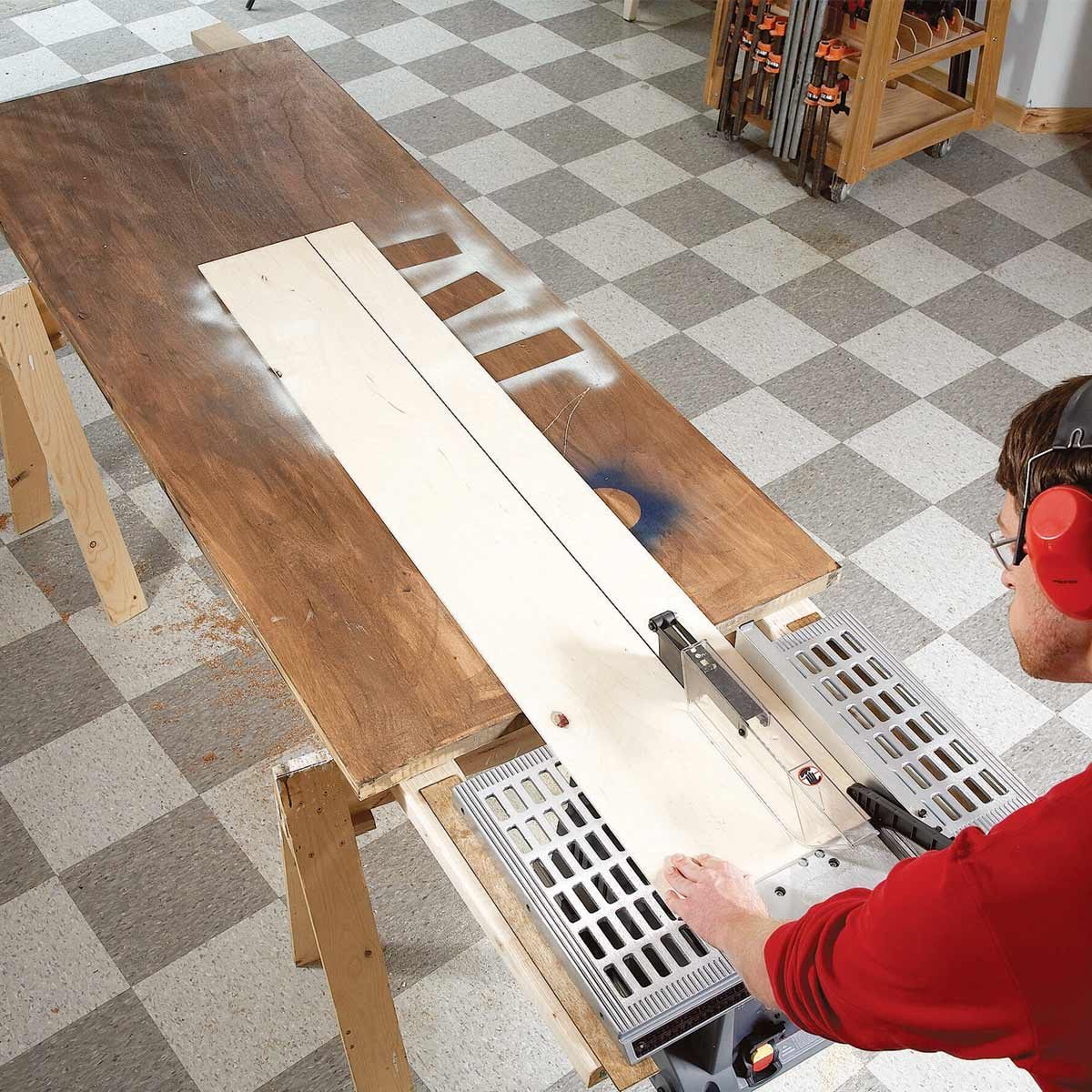 A person wearing ear protection cuts a wooden board on a table with a circular saw, surrounded by a workshop environment with checkered flooring.
