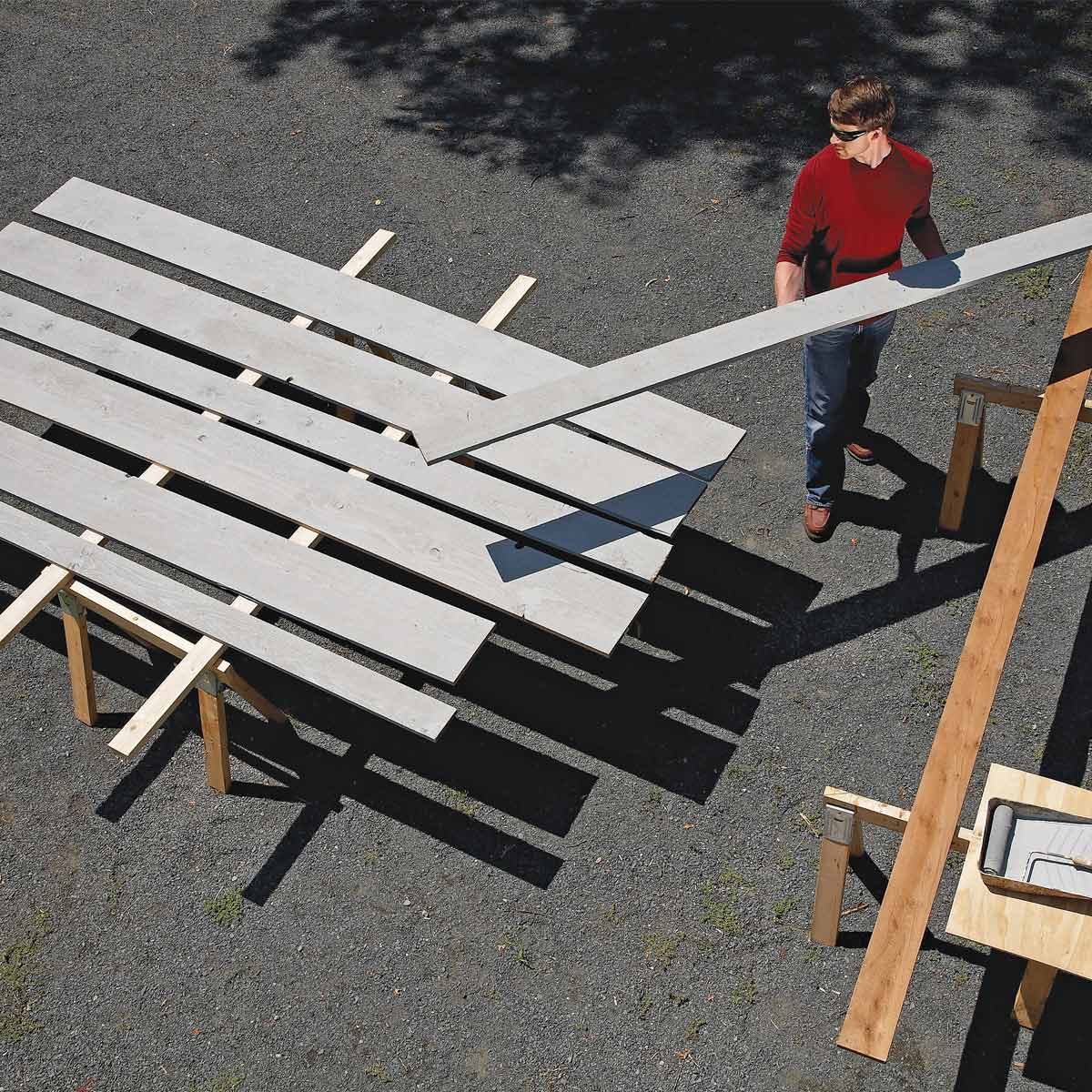 A man lifts a wooden plank from a stack on a construction site, surrounded by other planks and tools on a gravel surface.