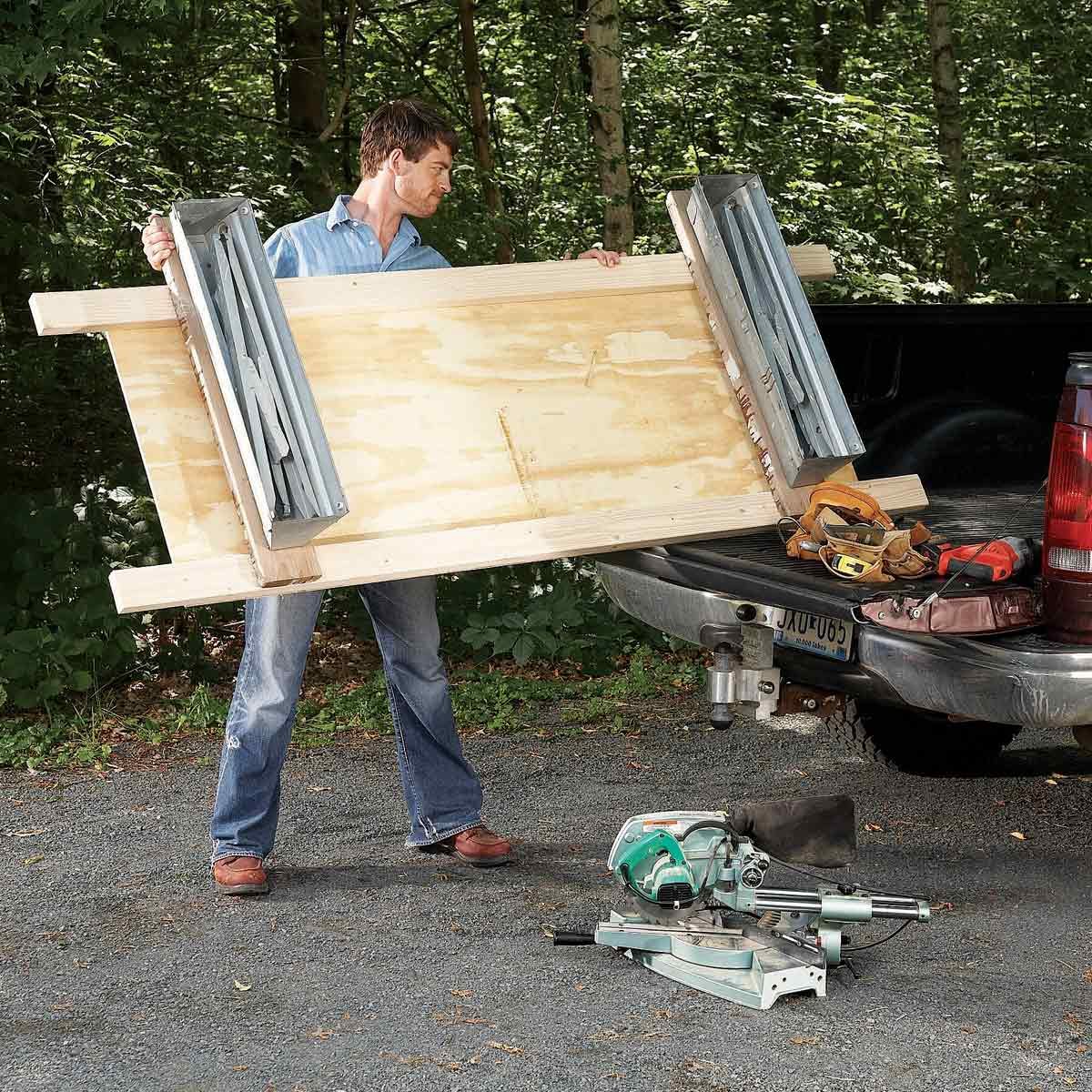 A man lifts a large wooden board from the back of a truck in a forested area, with a miter saw and tools on the ground nearby.