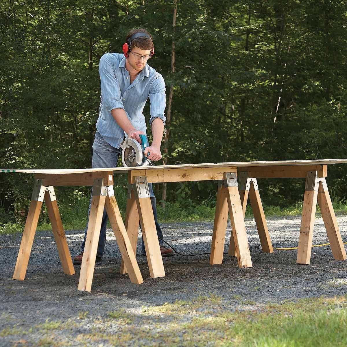 A man uses a circular saw to cut a wooden board, positioned on sawhorses outdoors amid a forested background.