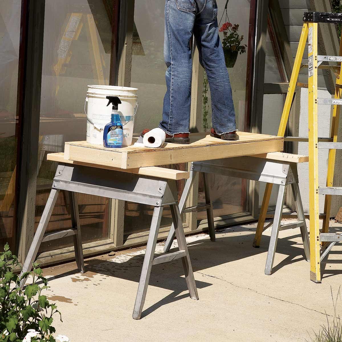 A person stands on a wooden platform supported by sawhorses, using cleaning supplies to work on a window in a sunny outdoor space.