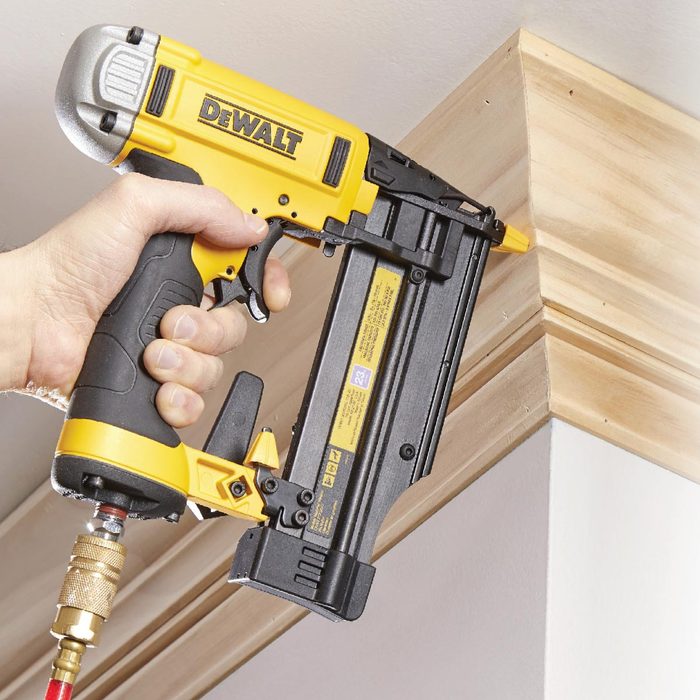 A hand holds a yellow and black nail gun, driving a nail into wooden trim at the corner of a wall and ceiling.