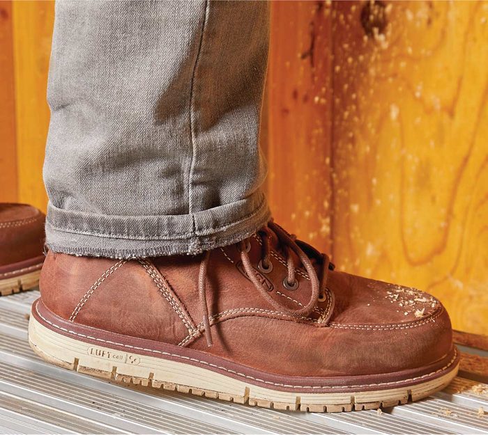 A brown leather boot stands on a metallic step, paired with rolled-up gray jeans, against a backdrop of wooden planks. Dust and wear are visible.