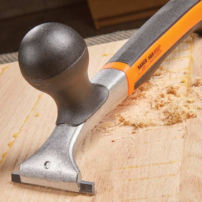 A scraper tool with a rounded handle rests on a wooden surface, surrounded by wood shavings, indicating recent use for smoothing or shaping wood.