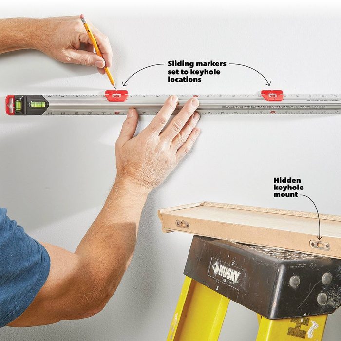 A person uses a level to mark keyhole locations on a wall, while a ladder supports an object with hidden keyhole mounts nearby.