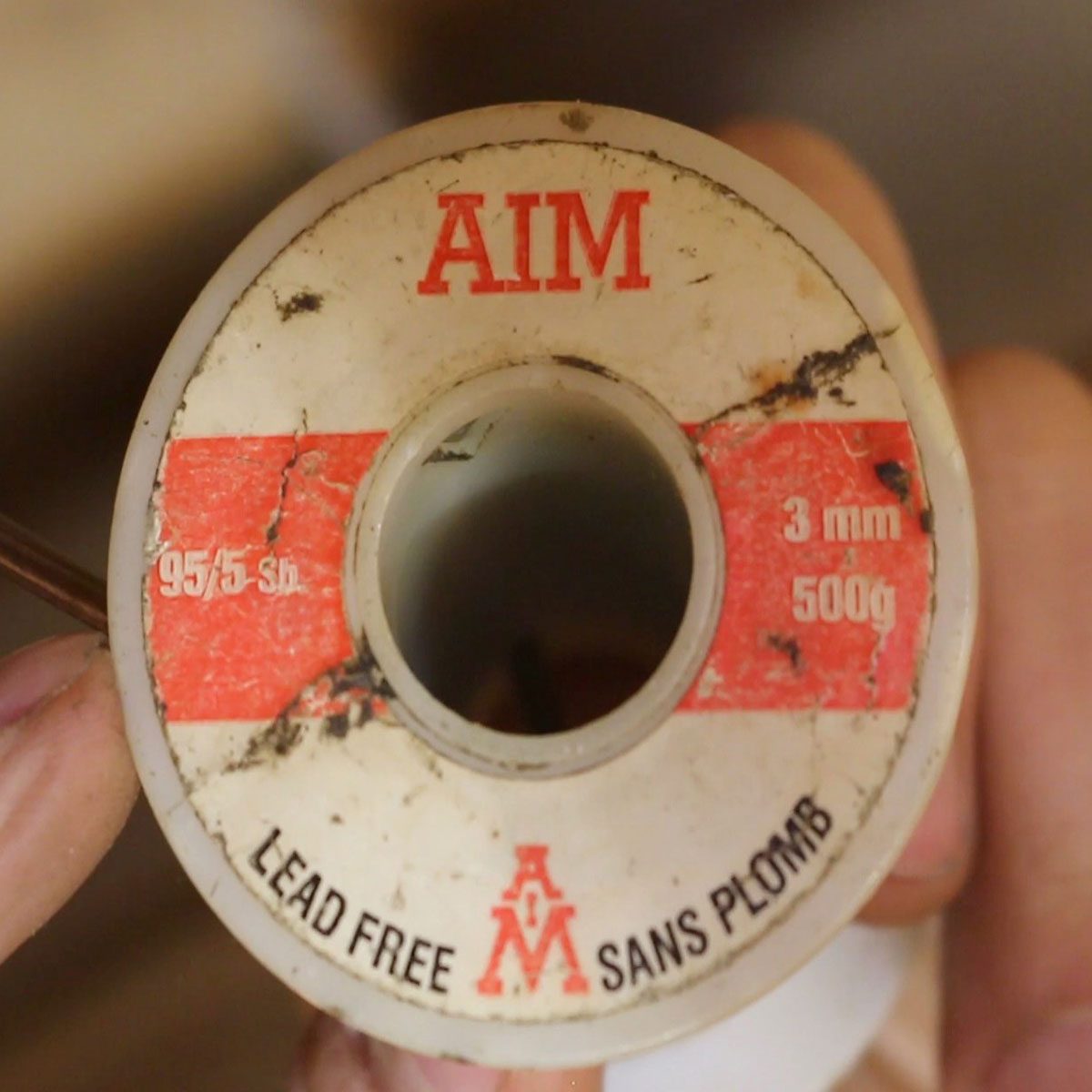 A spool of solder is held up, showing labels indicating specifications: "AIM," "LEAD FREE," "95/5 sb," "3 mm," and "500g." The surface appears worn.