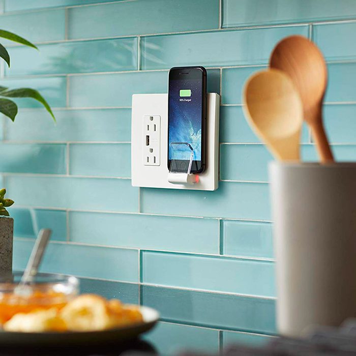 A smartphone is charging on a wall-mounted dock near a countertop, surrounded by kitchen utensils and decorative plants against a turquoise tiled wall.