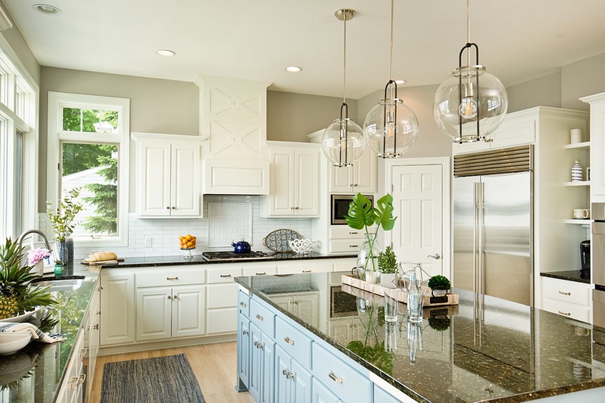 A modern kitchen features a central island with a dark countertop, surrounded by white cabinets, pendant lights, and decorative plants on the island.