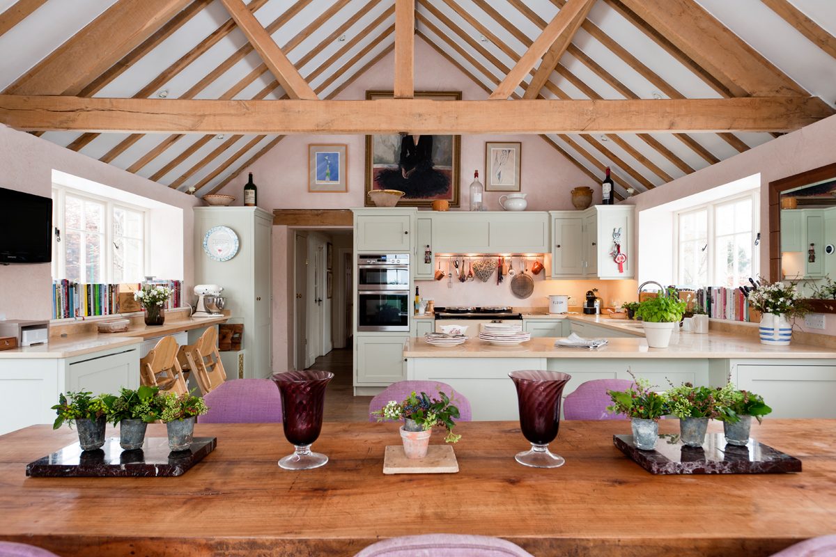 A wooden dining table is set with vases and plants, while a light, airy kitchen features storage, appliances, and decorative artwork under a vaulted ceiling.
