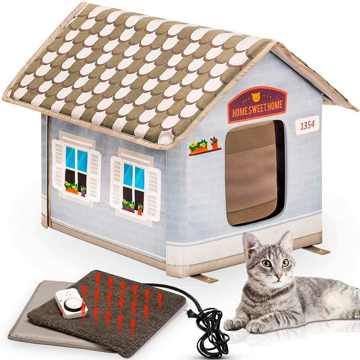 A plush cat house sits beside a heating pad, while a gray cat rests nearby. The house features a pitched roof and decorative windows.