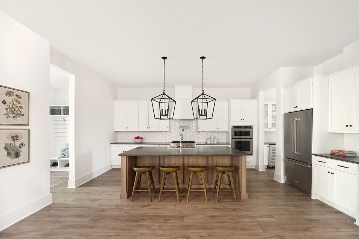 A kitchen features a central island with four wooden stools, two pendant lights hanging above, and modern appliances against white cabinetry in a bright environment.