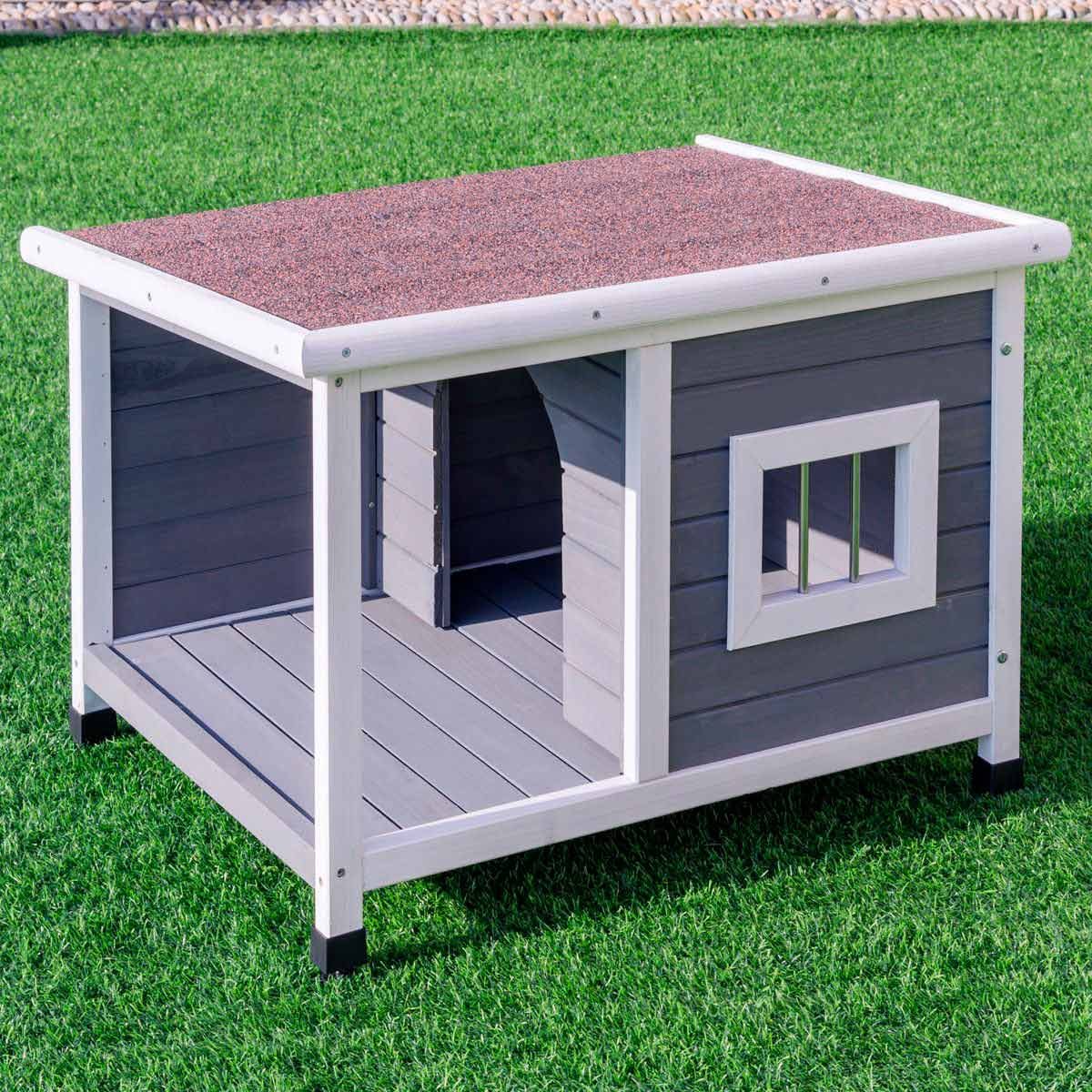 A pet house with a sloped, shingled roof sits on green grass. It has an open entrance, small window, and wooden panels.