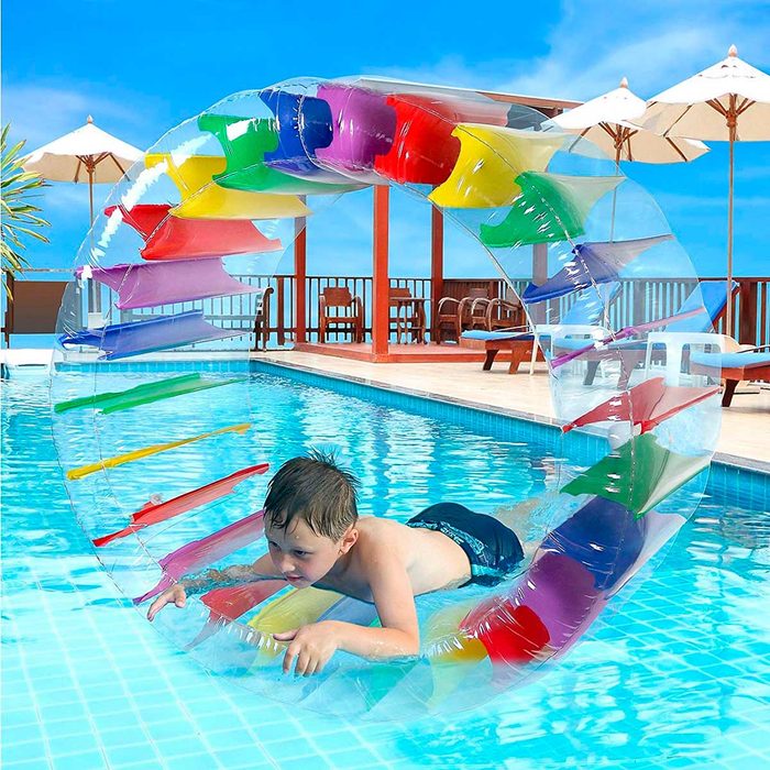 A child lies on a colorful inflatable water wheel in a swimming pool, surrounded by sun loungers and umbrellas under a clear blue sky.
