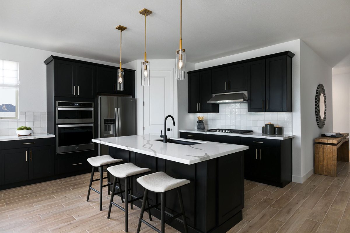 A modern kitchen features black cabinets and stainless appliances. A white countertop serves as a workspace, while pendant lights illuminate three bar stools nearby.