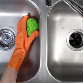 A hand wearing an orange glove scrubs a green sponge against a stainless steel sink, while the other sink basin remains empty.