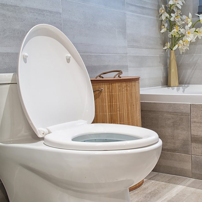 A white toilet with the lid lifted sits in a neatly tiled bathroom next to a bamboo basket and a bathtub, adorned with flowers.