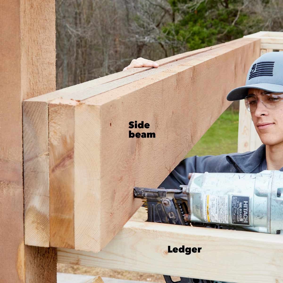 A person positions a large wooden side beam using a nail gun to secure it to a ledger, set against a natural outdoor backdrop.