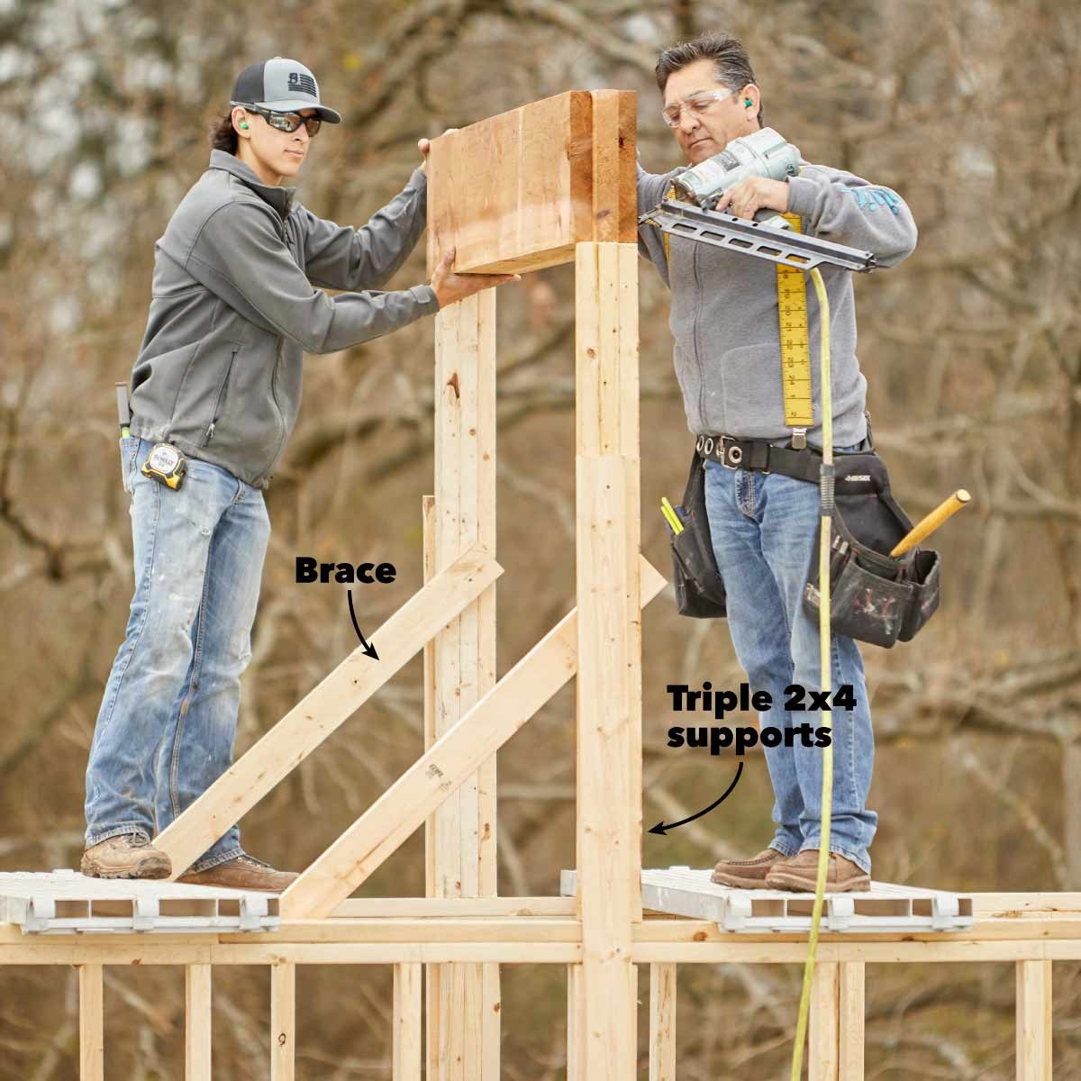 Two workers stand on a wooden structure, securing a large beam. They use tools for construction amidst a wooded outdoor setting.