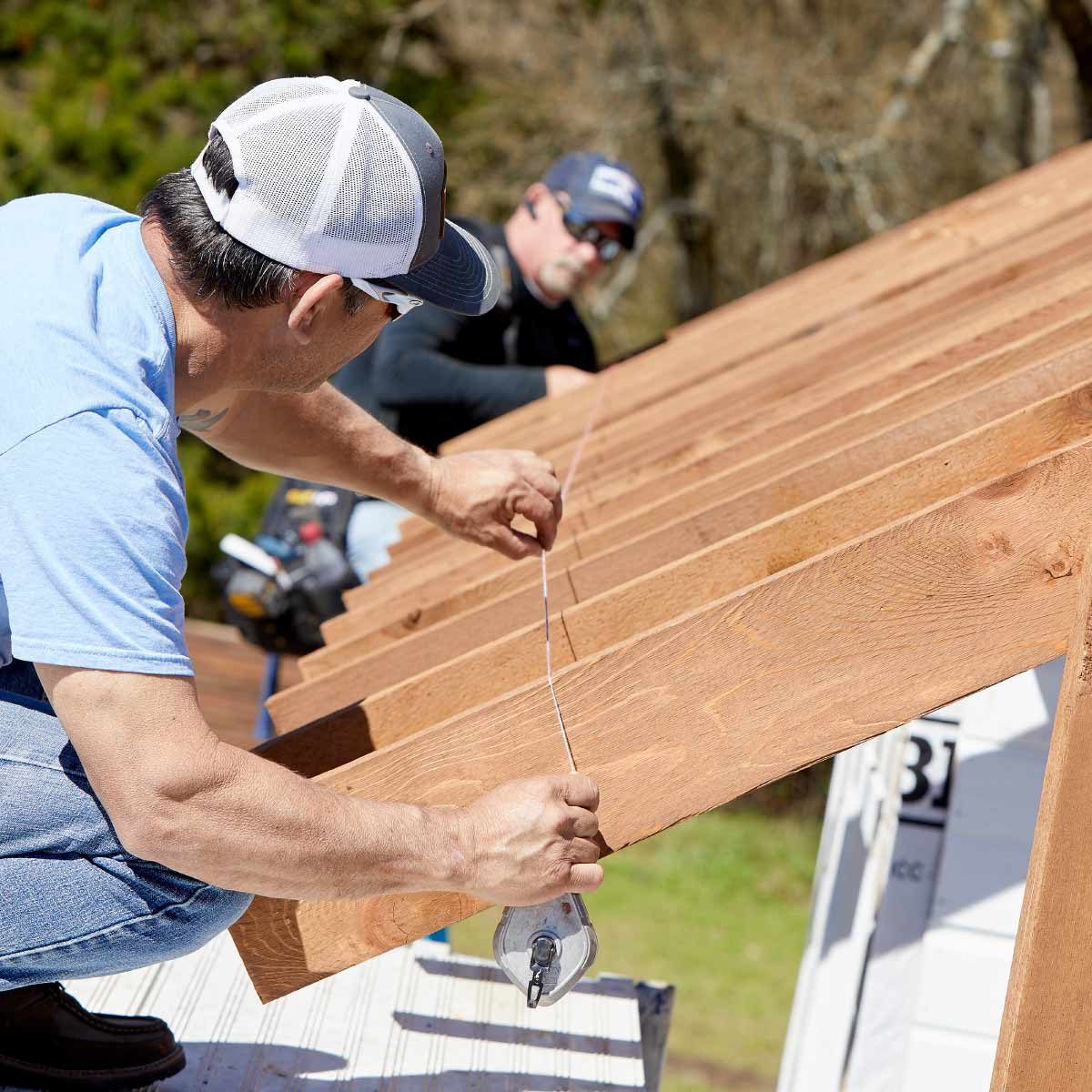 A man adjusts a measuring line while another stands nearby, both working on wooden rafters on a sunny day amid greenery.