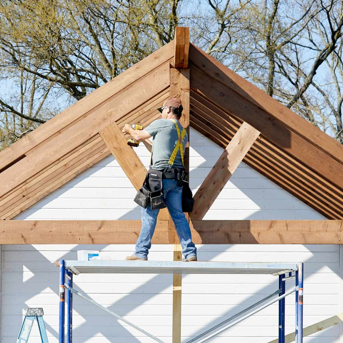 A construction worker uses a power drill to secure wooden beams atop a scaffold, with a partially completed building and trees visible in the background.