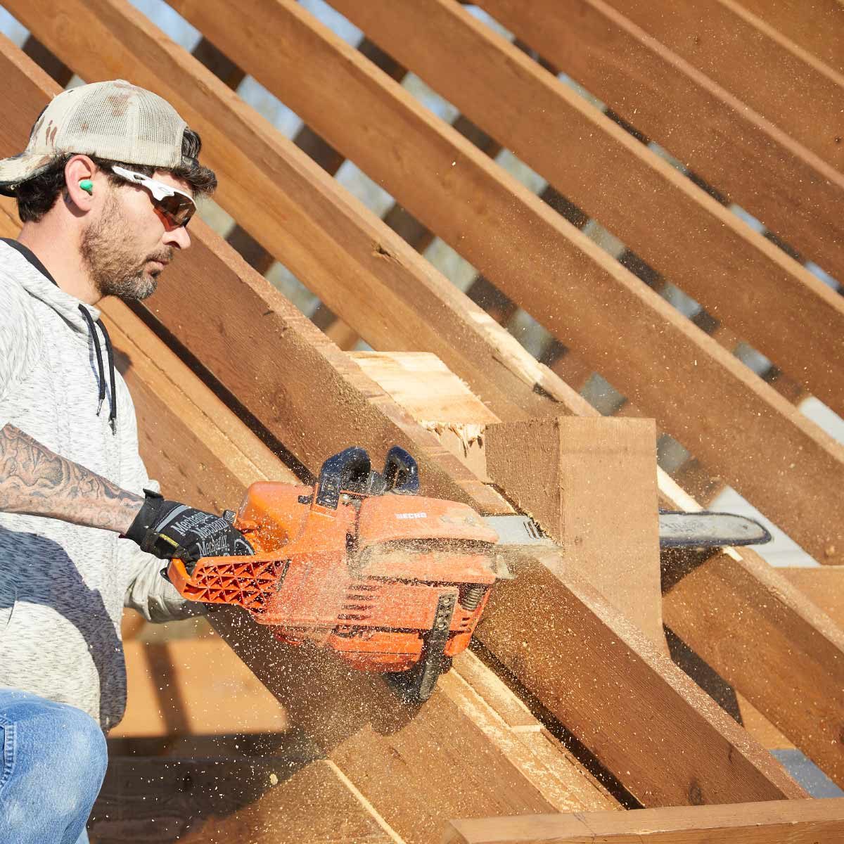 A worker uses a chainsaw to cut timber beams, creating sawdust that fills the air, surrounded by wooden structure rafters and bright sunlight.