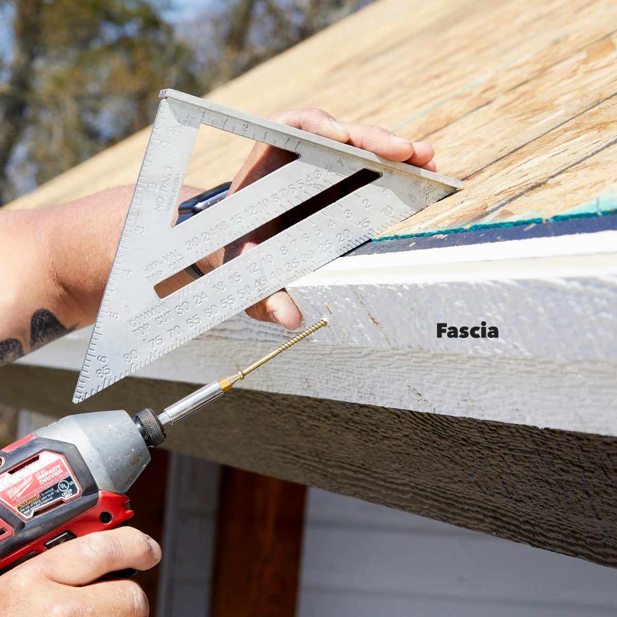 A person uses a square tool to align a drill while attaching a screw to a fascia board on a roof, surrounded by trees and a building.