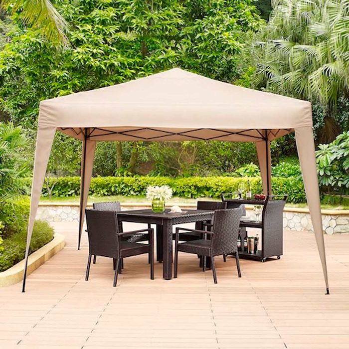 A beige canopy stands over a black table and four chairs in a lush garden, surrounded by greenery and wooden decking.