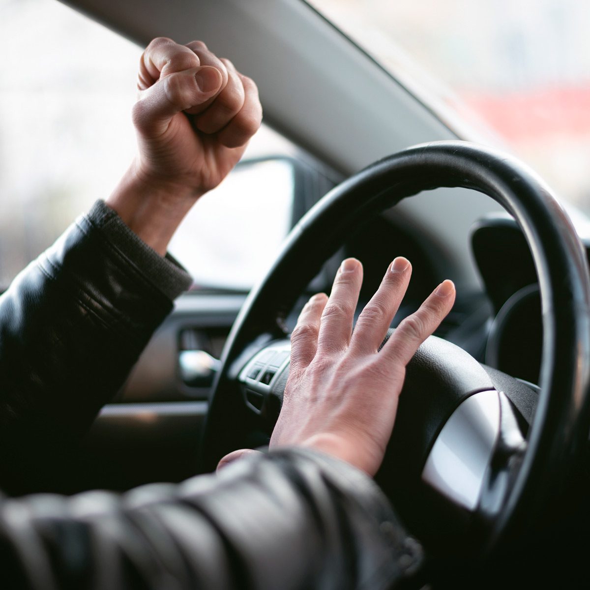 A hand grips a steering wheel while another hand is raised in frustration, indicating irritation inside a car, likely in a traffic situation.