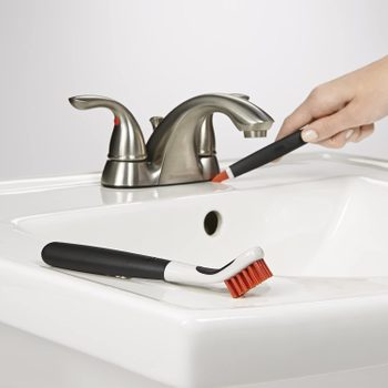 A hand holds a cleaning brush, scrubbing a sink near a metallic faucet, emphasizing a cleaning action in a bathroom setting.