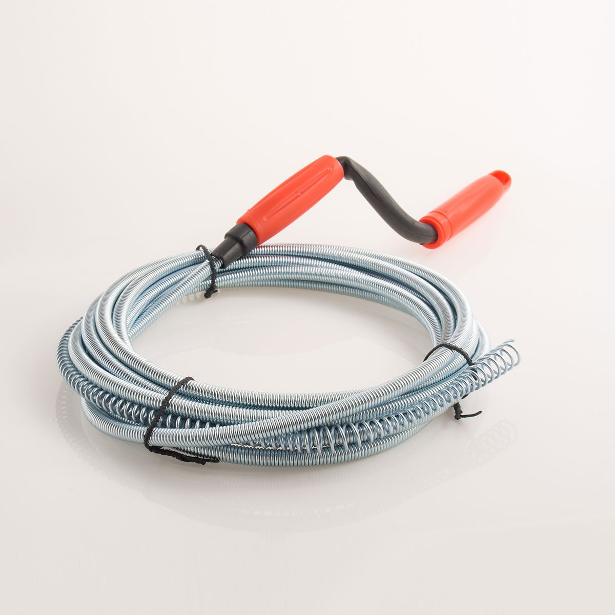 A coiled metal plumbing snake with a red handle lies on a reflective surface, designed for unclogging drains in pipes.