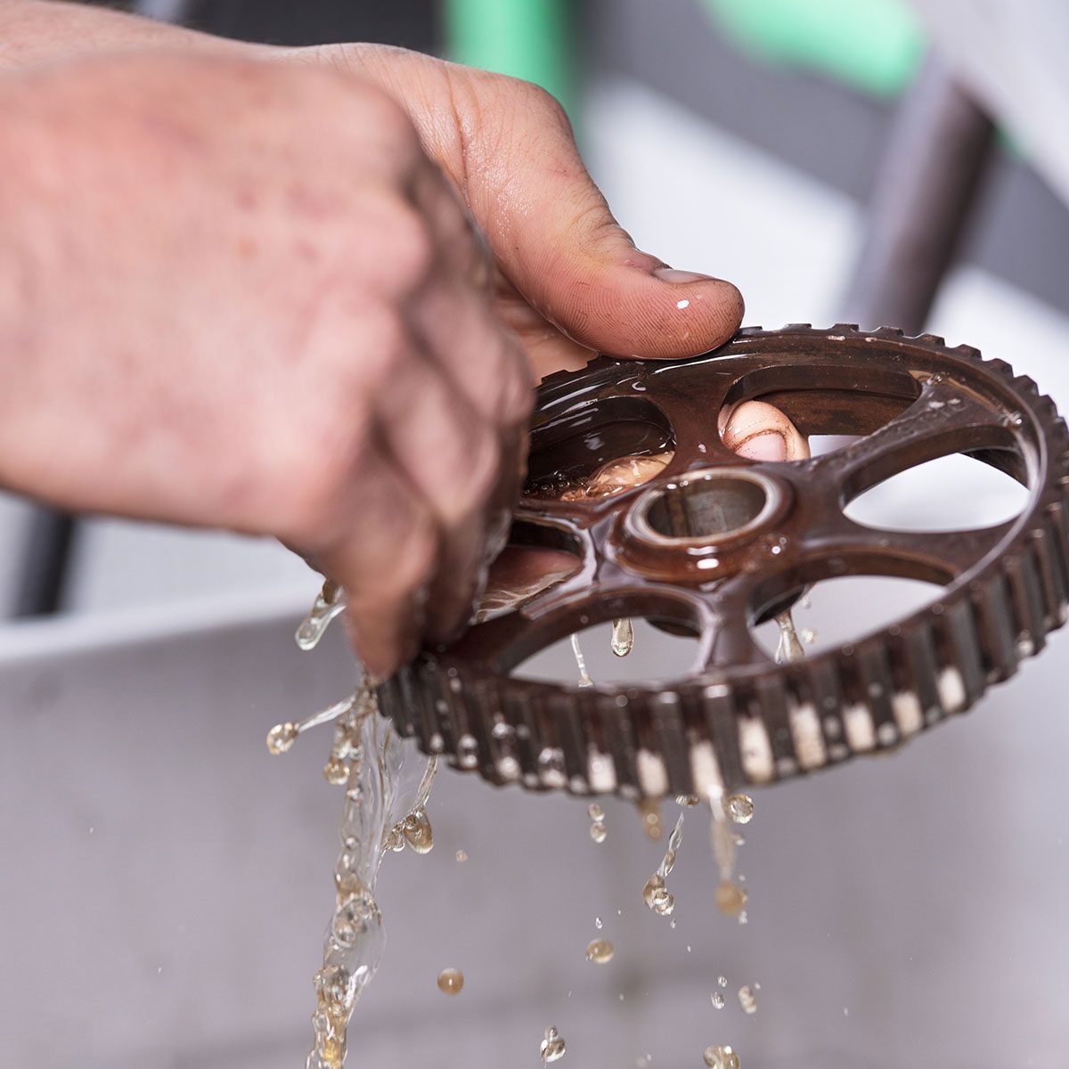 A hand holds a brown gear above a basin, water dripping off as it is rinsed, surrounded by a workshop environment.