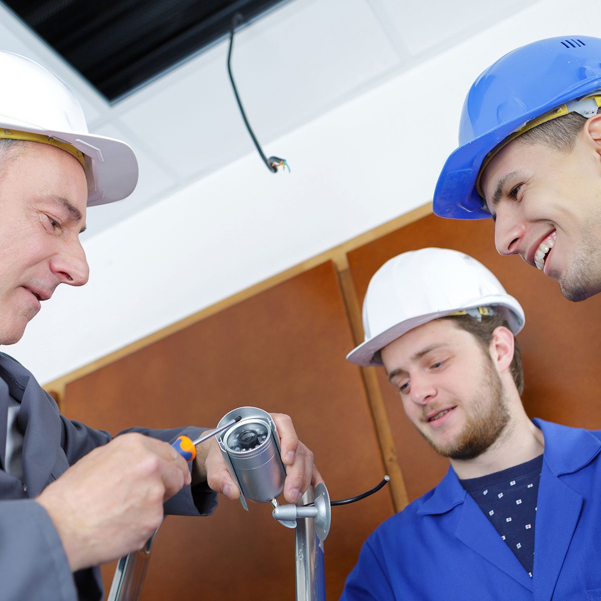 Two workers, wearing hard hats, install a security camera. One uses a screwdriver while the other observes, in a setting with wooden panels and overhead wiring.