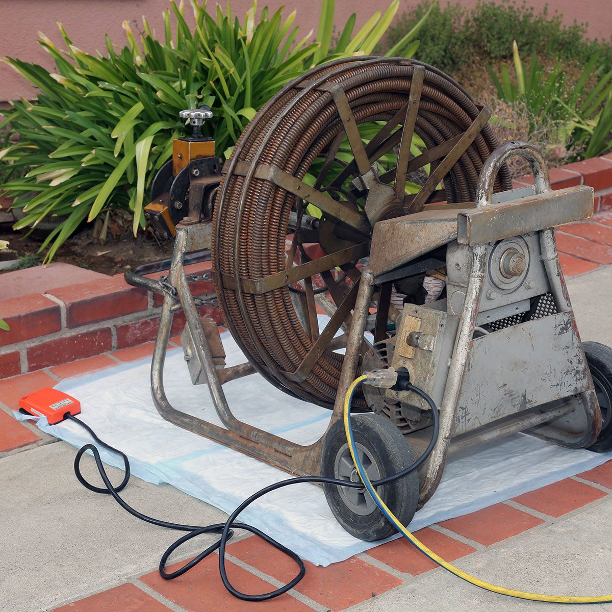 A large, metal drain auger stands on a protective sheet, with coiled cable and power cords leading to it, surrounded by greenery and brick pavement.