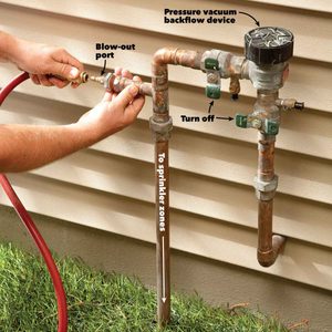 A hand connects a hose to a pipe with various fittings attached, against a house's siding, indicating maintenance for a sprinkler system.