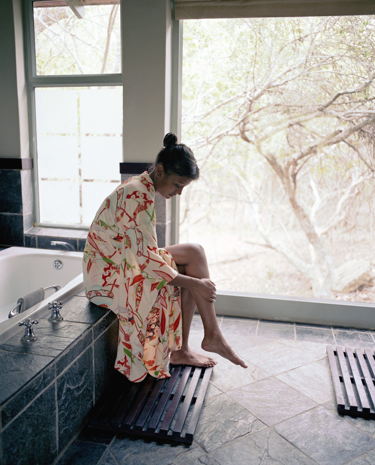 A person wearing a patterned robe sits on a stone ledge, reflecting thoughtfully, with large windows revealing a serene, nature-filled exterior.