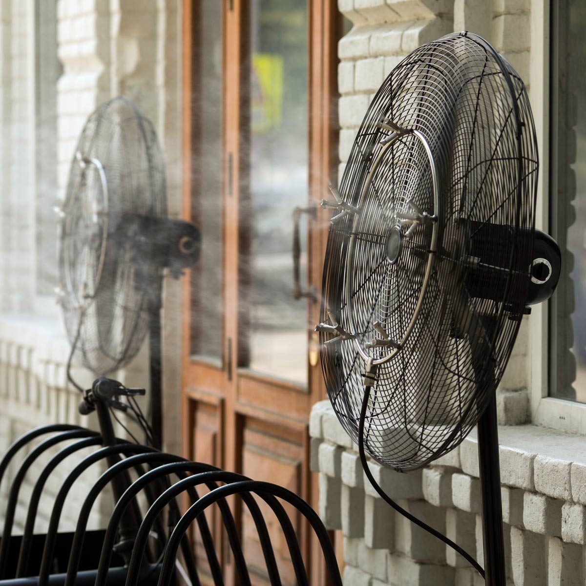 Two large fans are emitting mist while positioned near a set of double wooden doors, surrounded by a row of black metal chairs.