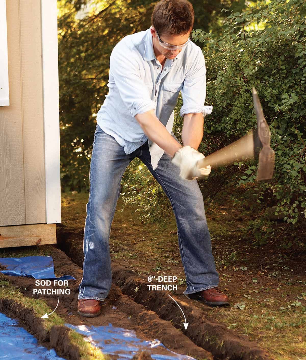 A man is digging an 8-inch deep trench in a yard using a pickaxe, surrounded by greenery and a blue tarp on the ground.
