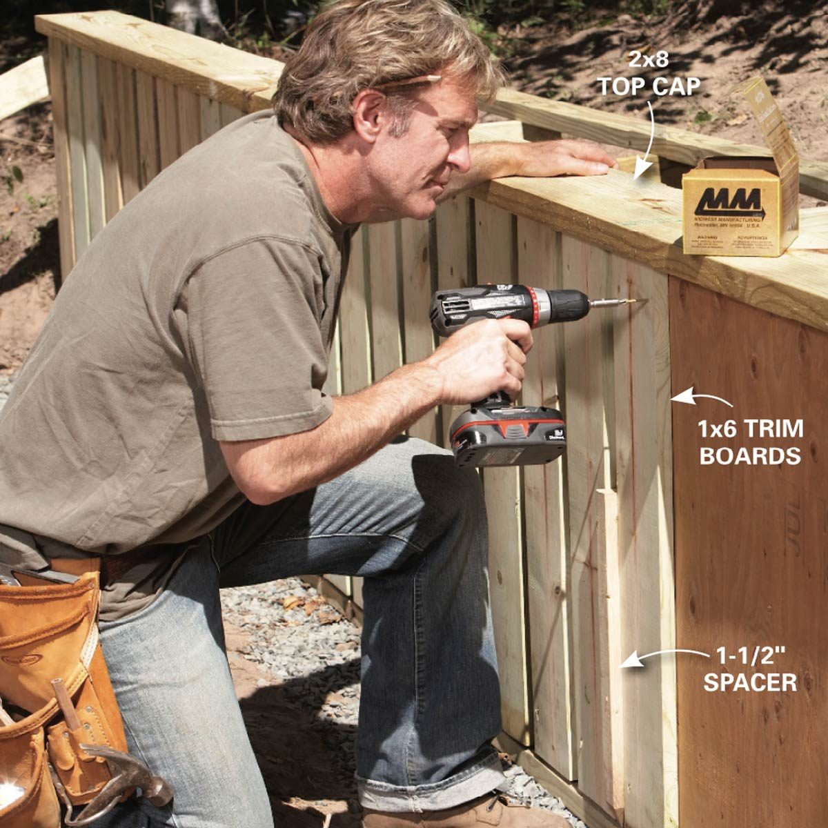A man kneels and uses a power drill to attach trim boards to a wooden structure while surrounded by gravel and partially completed woodwork.